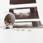 A white pill bottle lies on its side, spilling round beige tablets onto a white surface. Behind it, several blurred ultrasound images hint at the risks and complications associated with drug-induced abortion in this clinical, medical scene.
