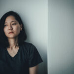 A woman with dark hair wearing a dark t-shirt sits against a light-colored wall, looking downcast and thoughtful, perhaps reflecting on the psychological reactions many women experience after an abortion, her face set in a neutral expression.