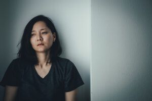 A woman with dark hair wearing a dark t-shirt sits against a light-colored wall, looking downcast and thoughtful, perhaps reflecting on the psychological reactions many women experience after an abortion, her face set in a neutral expression.