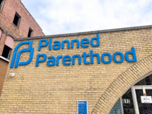 Exterior of a Planned Parenthood building at 612, featuring a large blue logo and signage on a tan brick wall, with an arched entrance and hours sign visible below. Highlighted in the 2024-25 Annual Report for its welcoming design.