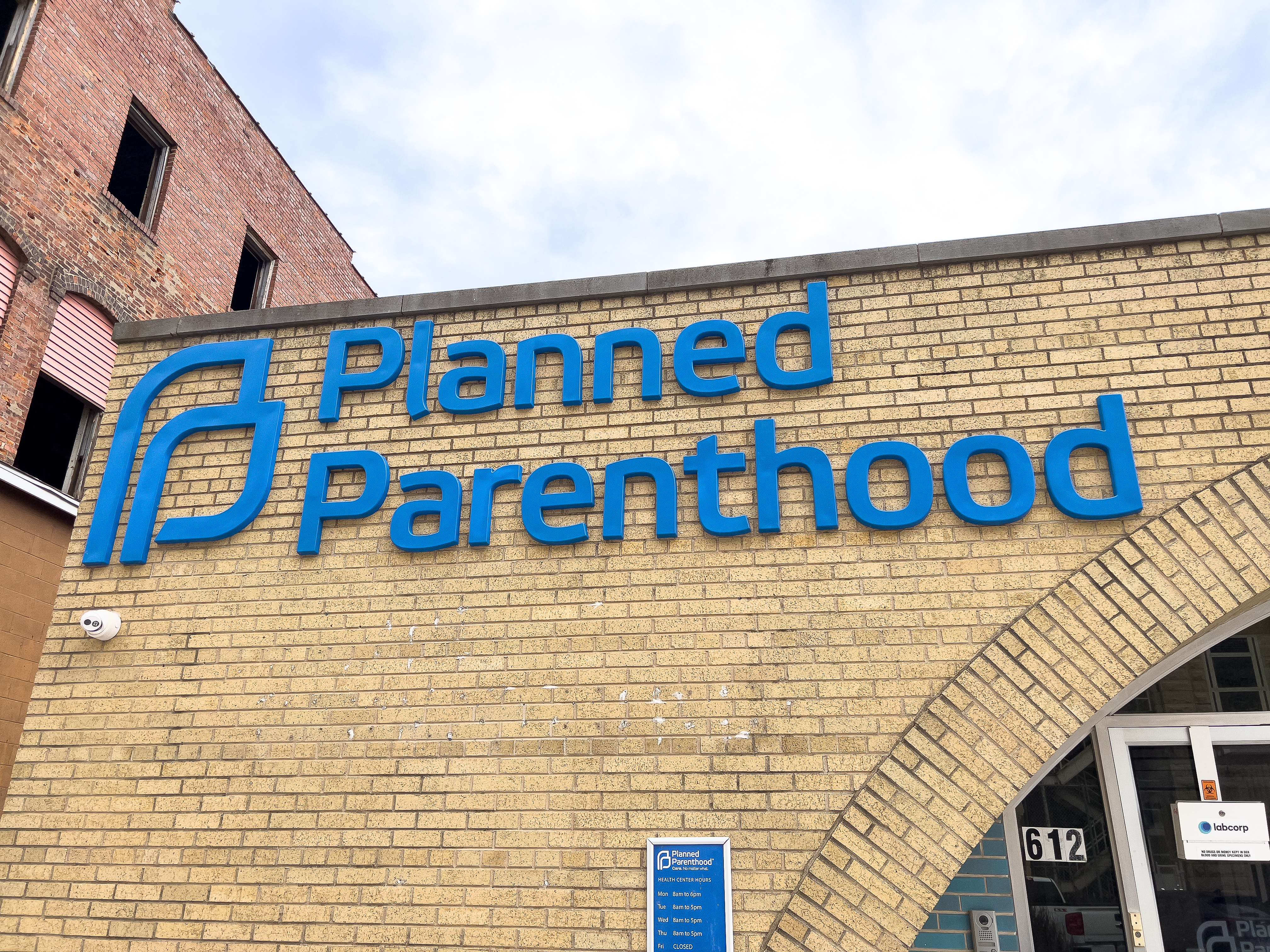 Exterior of a Planned Parenthood building at 612, featuring a large blue logo and signage on a tan brick wall, with an arched entrance and hours sign visible below. Highlighted in the 2024-25 Annual Report for its welcoming design.