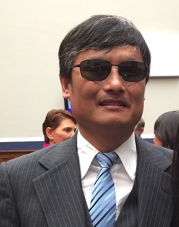 A man with short black hair wearing dark sunglasses, a striped suit, white shirt, and blue striped tie stands indoors. There are people and a wall in the background.