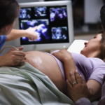 A pregnant woman lies on an exam table during an ultrasound. A medical professional points at images on a monitor, while another person sits beside the woman, touching her arm supportively