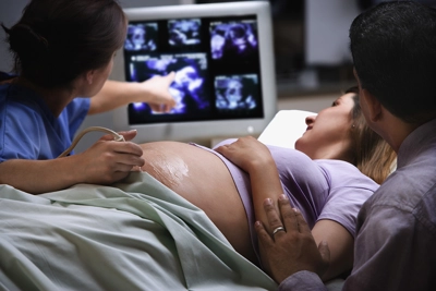A pregnant woman lies on an exam table during an ultrasound. A medical professional points at images on a monitor, while another person sits beside the woman, touching her arm supportively