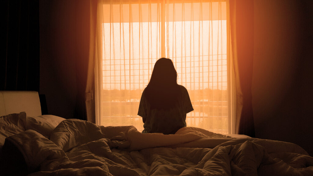 A person sits on an unmade bed facing a bright window with sheer curtains, their silhouette outlined by the warm, orange glow of sunlight—evoking a moment of quiet reflection on abortion in the dimly lit room.