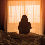 A person sits on an unmade bed facing a bright window with sheer curtains, their silhouette outlined by the warm, orange glow of sunlight—evoking a moment of quiet reflection on abortion in the dimly lit room.
