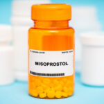 An orange prescription bottle labeled MISOPROSTOL, a common abortion pill, filled with yellow tablets sits in front of several white medicine bottles against a light blue background.