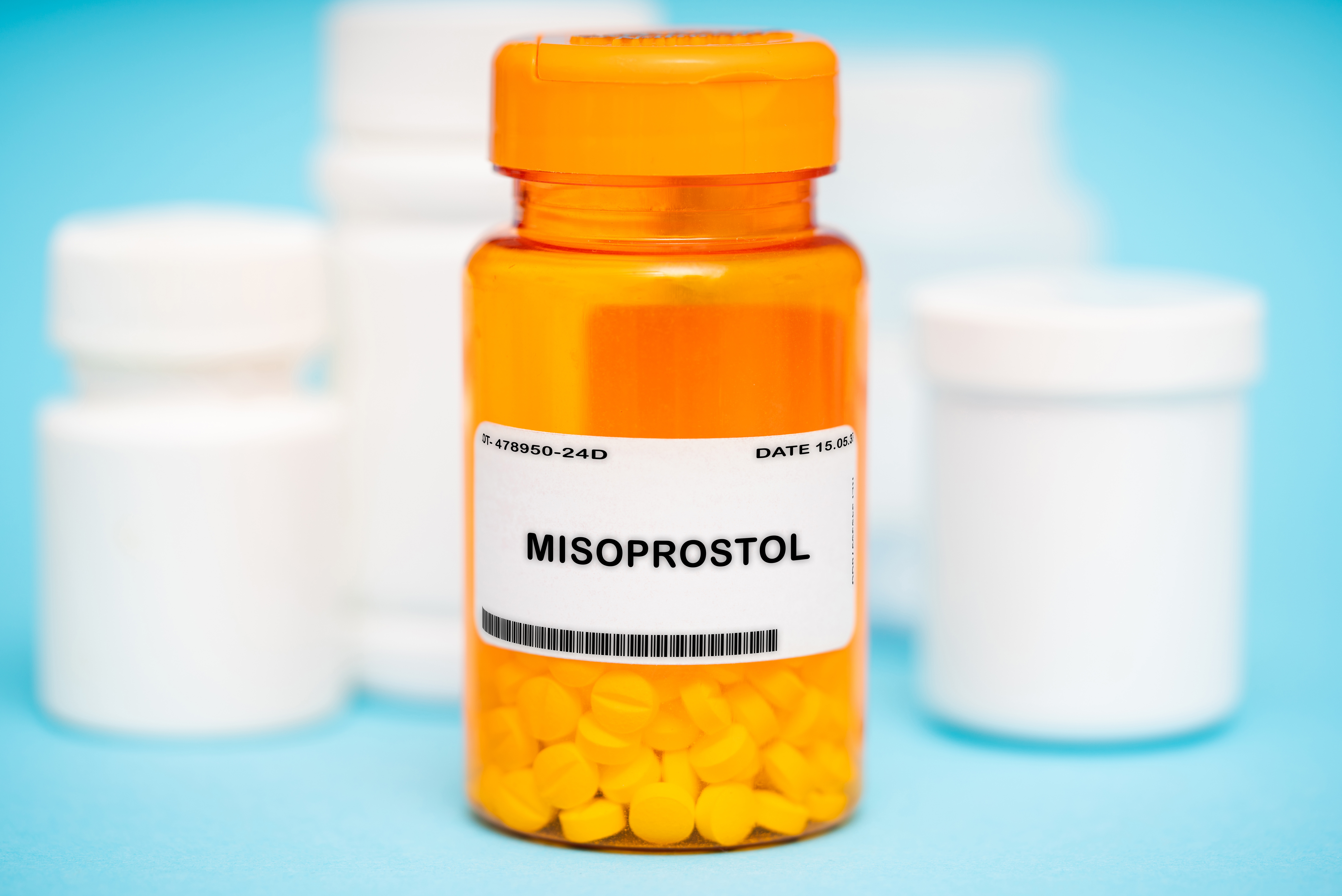 An orange prescription bottle labeled MISOPROSTOL, a common abortion pill, filled with yellow tablets sits in front of several white medicine bottles against a light blue background.
