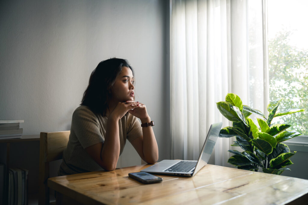 A woman sits at a wooden table with a laptop and phone, gazing thoughtfully out a window. Sunlight filters through sheer curtains, and a green potted plant is beside her as she researches online abortion drug access in post-Dobbs America.