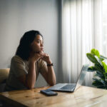 A woman sits at a wooden table with a laptop and phone, gazing thoughtfully out a window. Sunlight filters through sheer curtains, and a green potted plant is beside her as she researches online abortion drug access in post-Dobbs America.