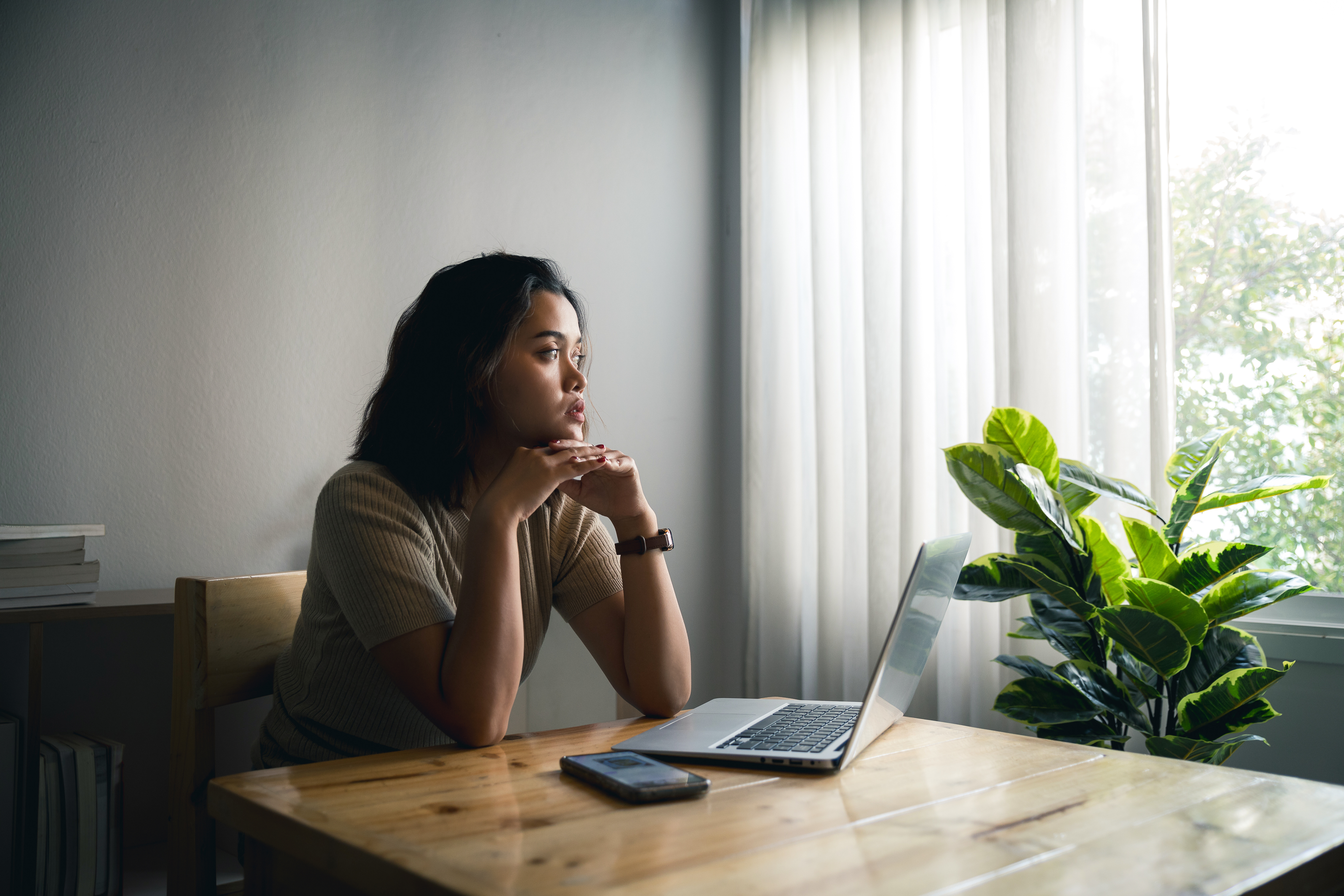 A woman sits at a wooden table with a laptop and phone, gazing thoughtfully out a window. Sunlight filters through sheer curtains, and a green potted plant is beside her as she researches online abortion drug access in post-Dobbs America.