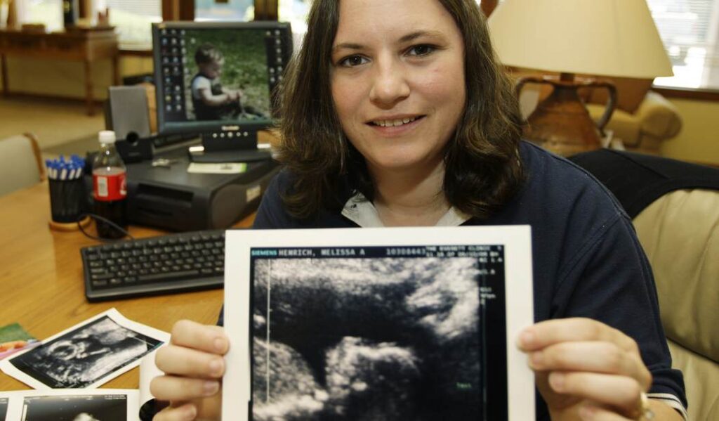 A woman smiles and holds up an ultrasound image of an unborn baby, seated at a desk with a computer, photos, and a drink bottle in the background.