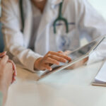 A doctor in a white coat uses a digital tablet to show information to a patient across a desk. The patient's hands are clasped.