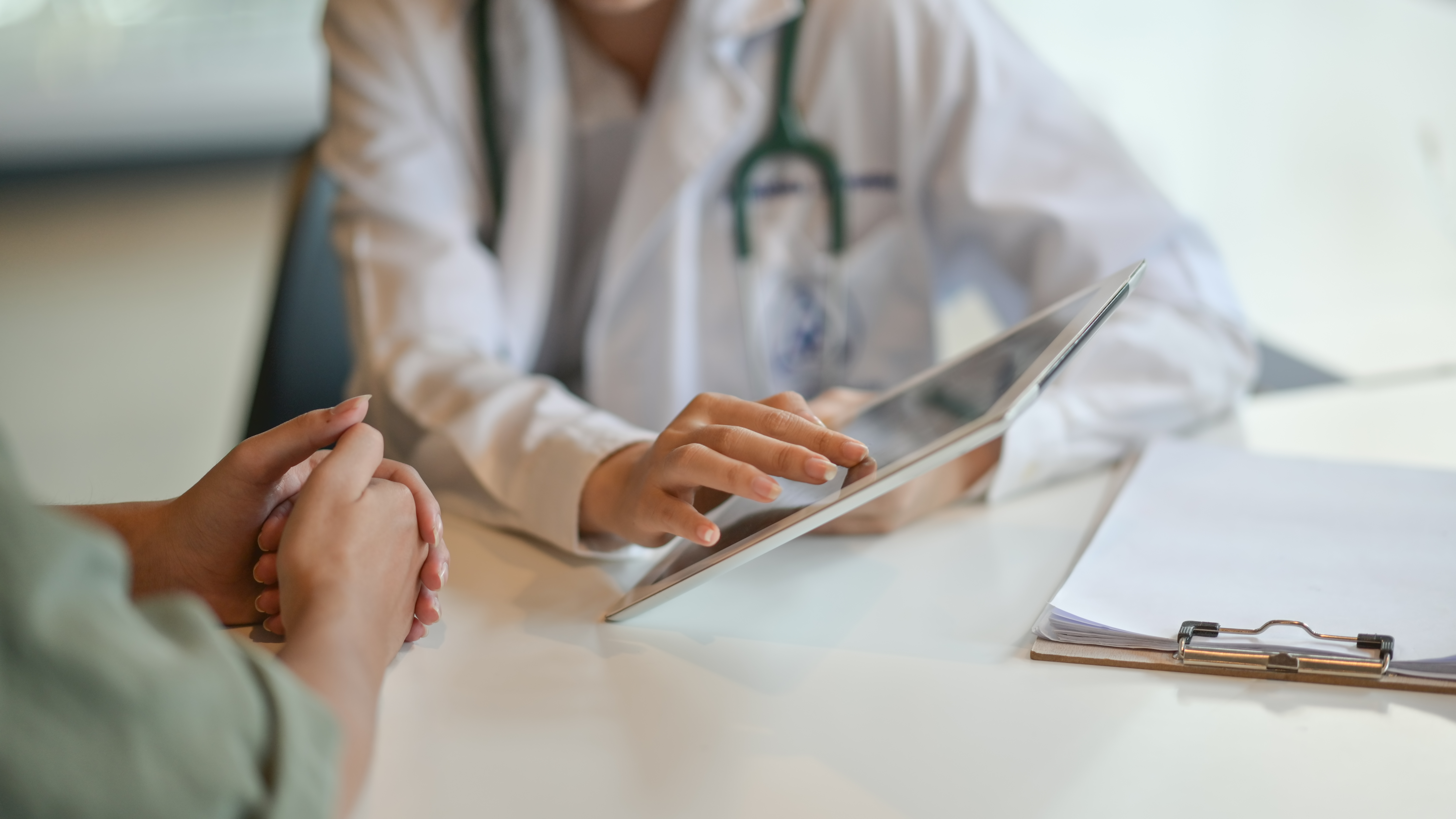 A doctor in a white coat uses a digital tablet to show information to a patient across a desk. The patient's hands are clasped.