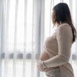 A pregnant woman stands indoors by a window with white curtains, gently holding her belly and looking outside. Natural light fills the room, reflecting hope for improving interventions for substance use disorder in pregnant women.