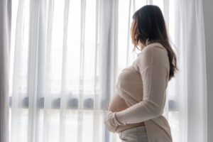 A pregnant woman stands indoors by a window with white curtains, gently holding her belly and looking outside. Natural light fills the room, reflecting hope for improving interventions for substance use disorder in pregnant women.