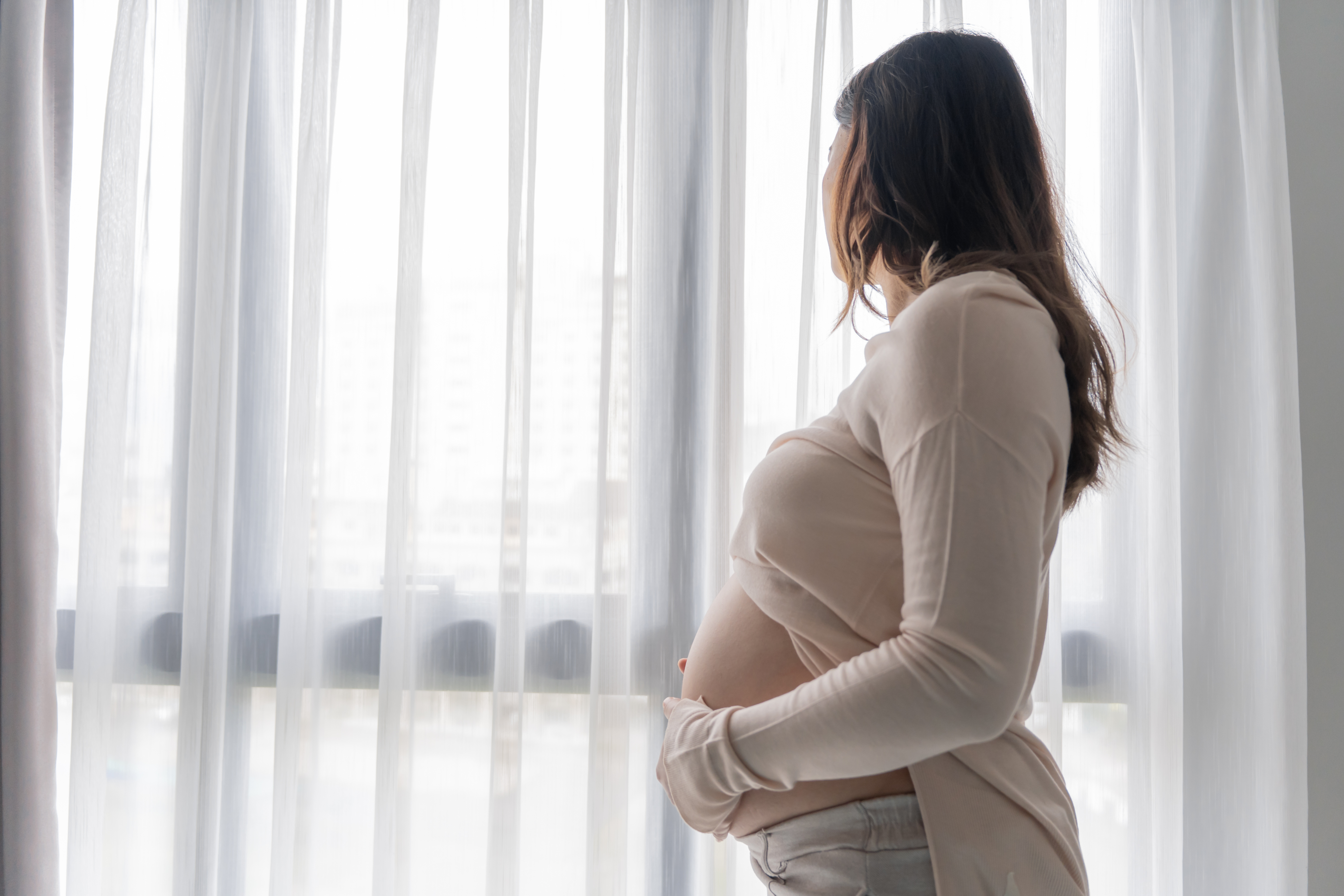 A pregnant woman stands indoors by a window with white curtains, gently holding her belly and looking outside. Natural light fills the room, reflecting hope for improving interventions for substance use disorder in pregnant women.