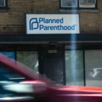 A red vehicle passes in front of a brick building with a Planned Parenthood sign above the entrance.