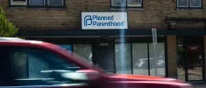 A red vehicle passes in front of a brick building with a Planned Parenthood sign above the entrance.