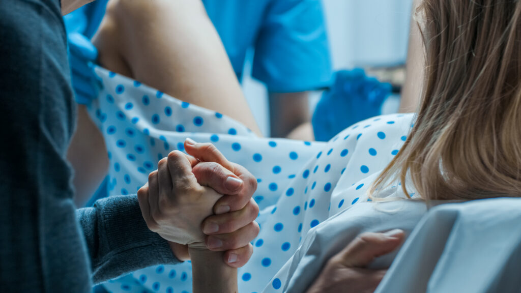 A woman in a hospital gown is in labor, holding someone's hand for support while medical staff assist during childbirth, closely monitoring for any medical indications that might require separating mother and unborn child.