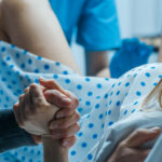 A woman in a hospital gown is in labor, holding someone's hand for support while medical staff assist during childbirth, closely monitoring for any medical indications that might require separating mother and unborn child.