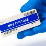 A hand in a blue glove holds a box labeled Mifepristone Tablets with a barcode and description stating its use for pregnancy termination and medical abortion, while recent controversies have brought increased attention to both Mifepristone and its uses beyond abortion.