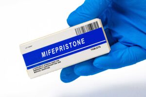 A hand in a blue glove holds a box labeled Mifepristone Tablets with a barcode and description stating its use for pregnancy termination and medical abortion, while recent controversies have brought increased attention to both Mifepristone and its uses beyond abortion.
