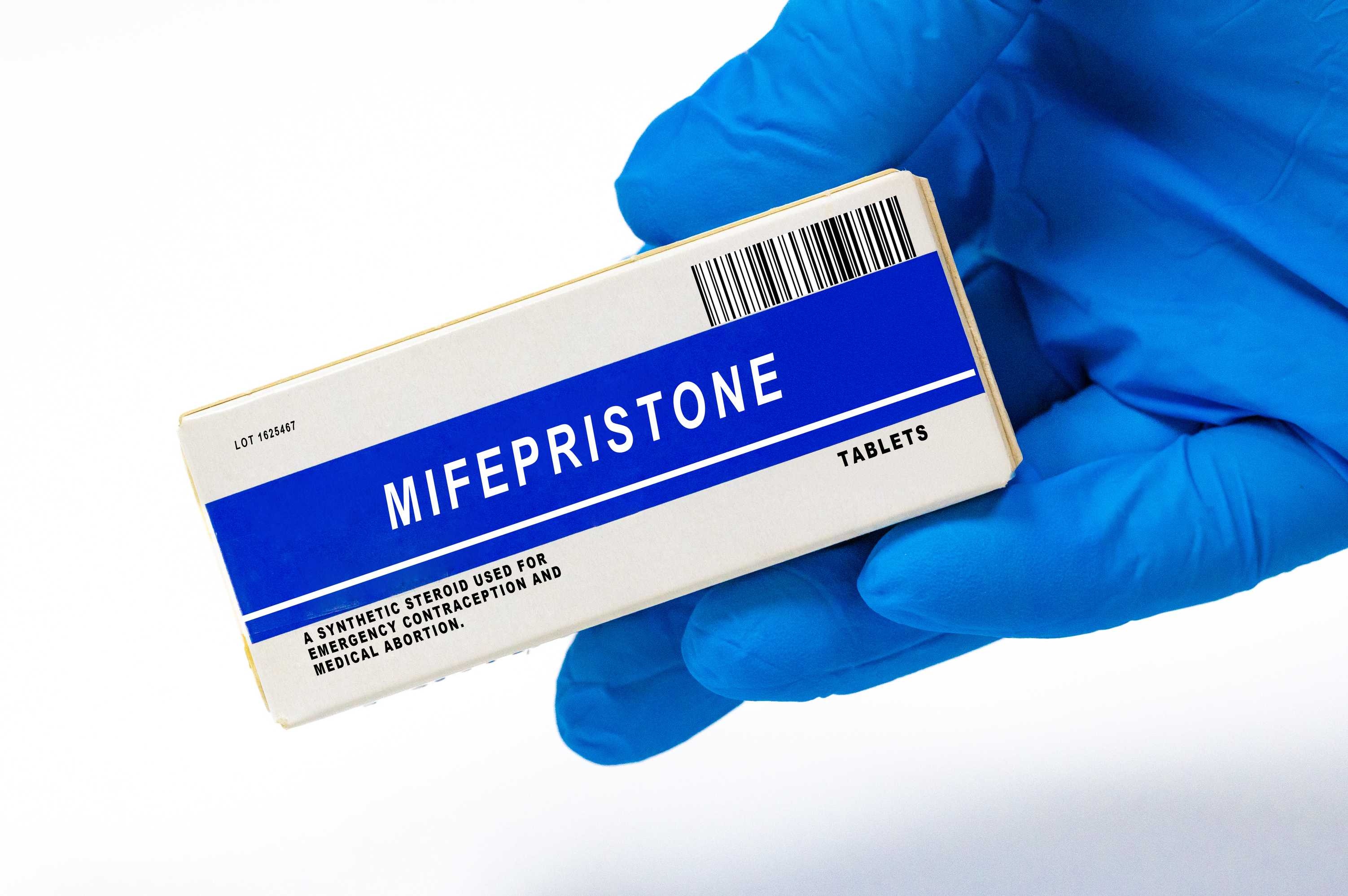 A hand in a blue glove holds a box labeled Mifepristone Tablets with a barcode and description stating its use for pregnancy termination and medical abortion, while recent controversies have brought increased attention to both Mifepristone and its uses beyond abortion.