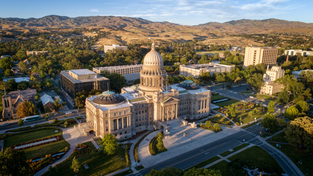 Aerial view of a grand capitol building with a central dome, surrounded by trees and city streets, with mountains in the background under a clear sky—the seat of the Idaho Board of Medicine, recently noted in cases such as Seyb v. Members of the Idaho Board of Medicine concerning debates over the federal right to abortion.