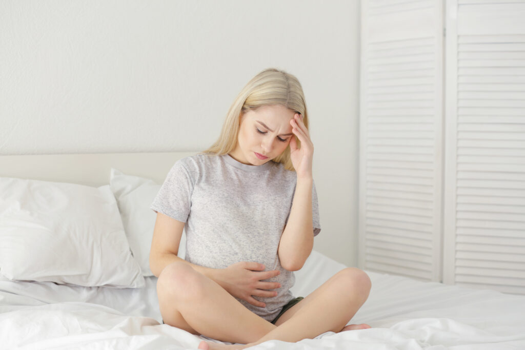 A woman sits cross-legged on a bed, holding her stomach with one hand and touching her forehead with the other, appearing to be in discomfort or pain—an image that may reflect the emotional and physical toll following pregnancy loss.