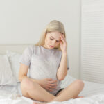 A woman sits cross-legged on a bed, holding her stomach with one hand and touching her forehead with the other, appearing to be in discomfort or pain—an image that may reflect the emotional and physical toll following pregnancy loss.