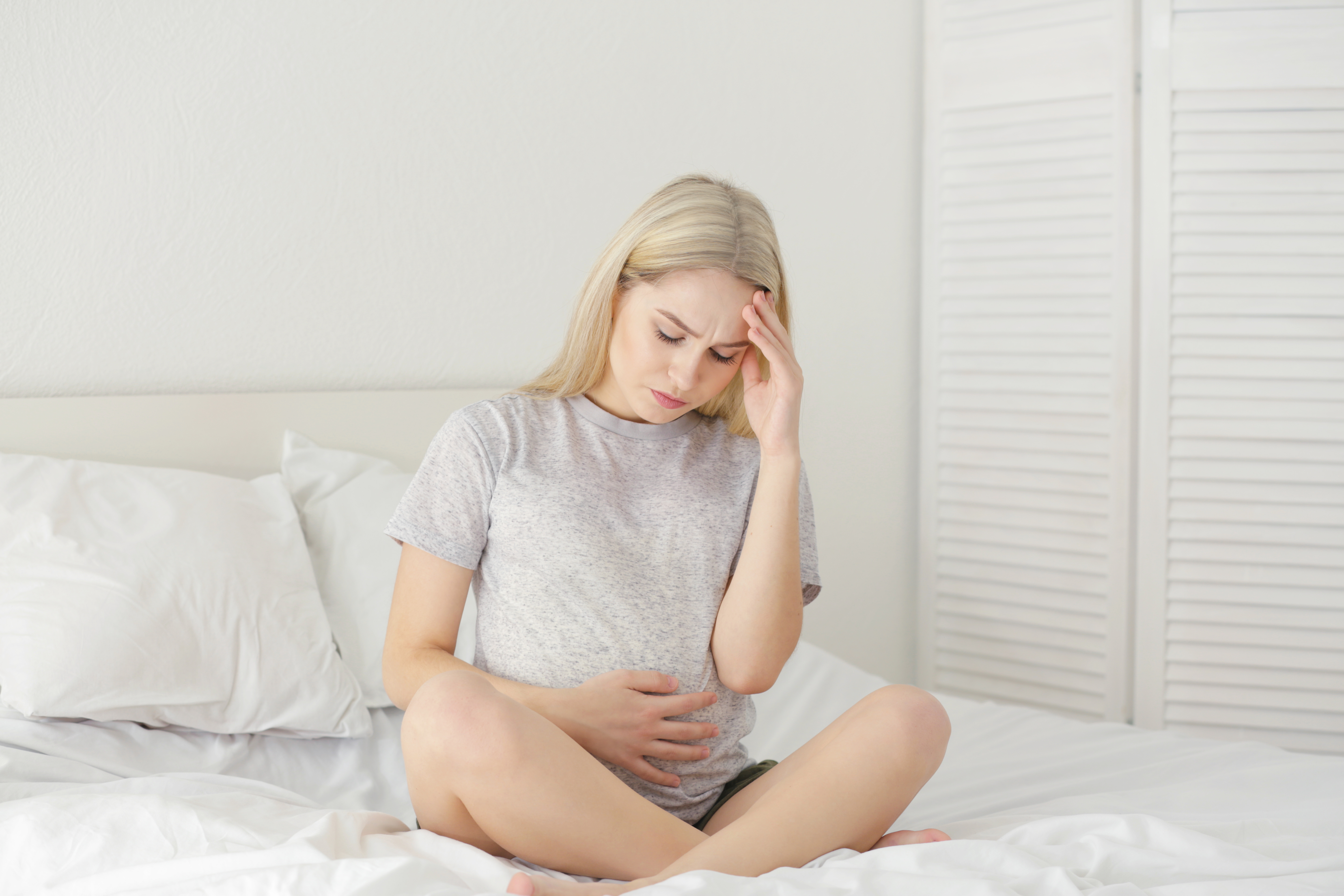 A woman sits cross-legged on a bed, holding her stomach with one hand and touching her forehead with the other, appearing to be in discomfort or pain—an image that may reflect the emotional and physical toll following pregnancy loss.