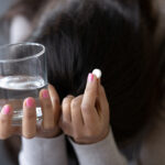 A person with pink nail polish holds a glass of water in one hand and a white pill in the other, their head bowed down, partially obscured by their hair—a quiet moment that reflects many women’s experiences as shared through online narratives about medication abortion.
