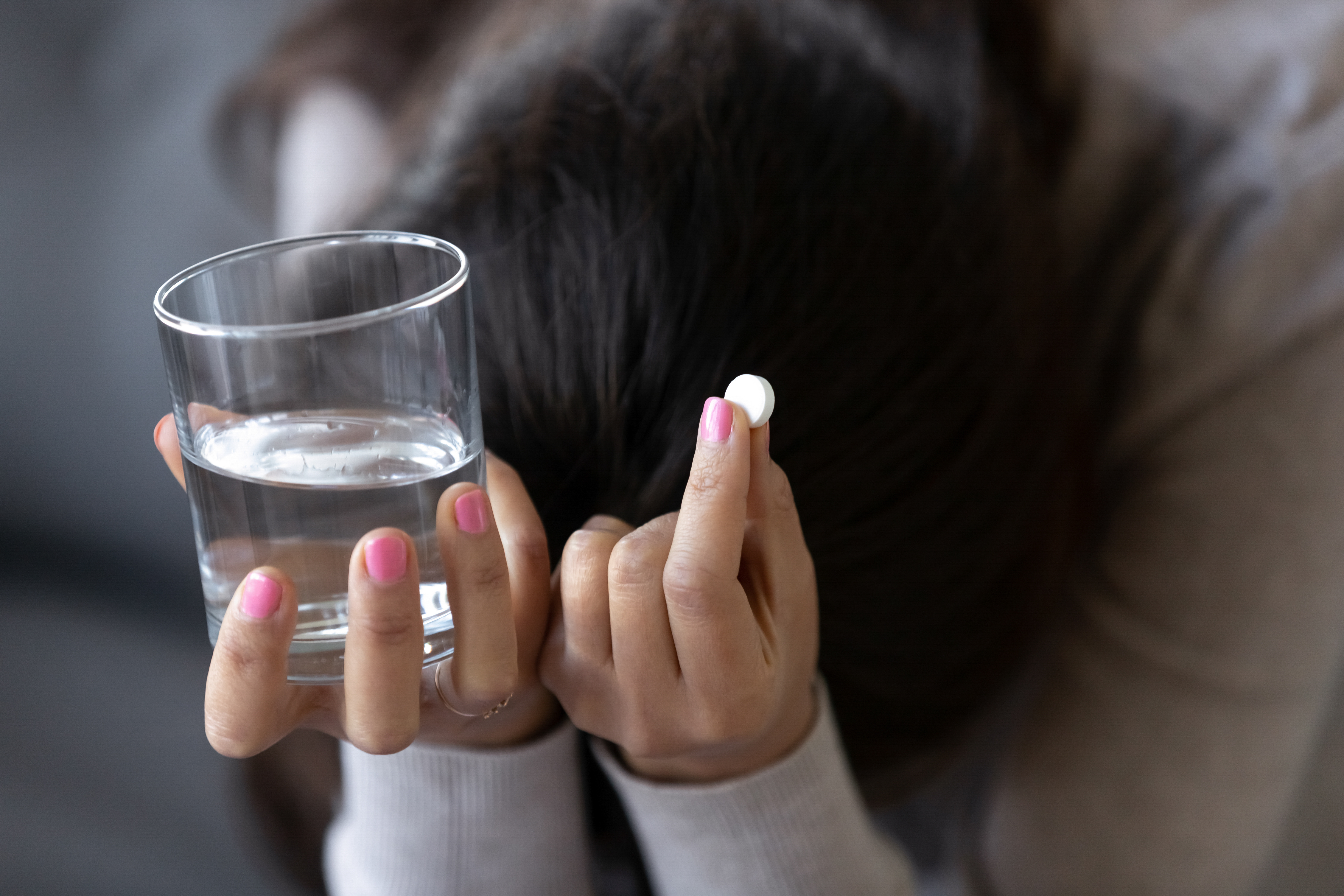 A person with pink nail polish holds a glass of water in one hand and a white pill in the other, their head bowed down, partially obscured by their hair—a quiet moment that reflects many women’s experiences as shared through online narratives about medication abortion.