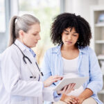 A doctor shows a tablet to a young woman patient sitting beside her in a medical office; the two appear to be discussing health information together.