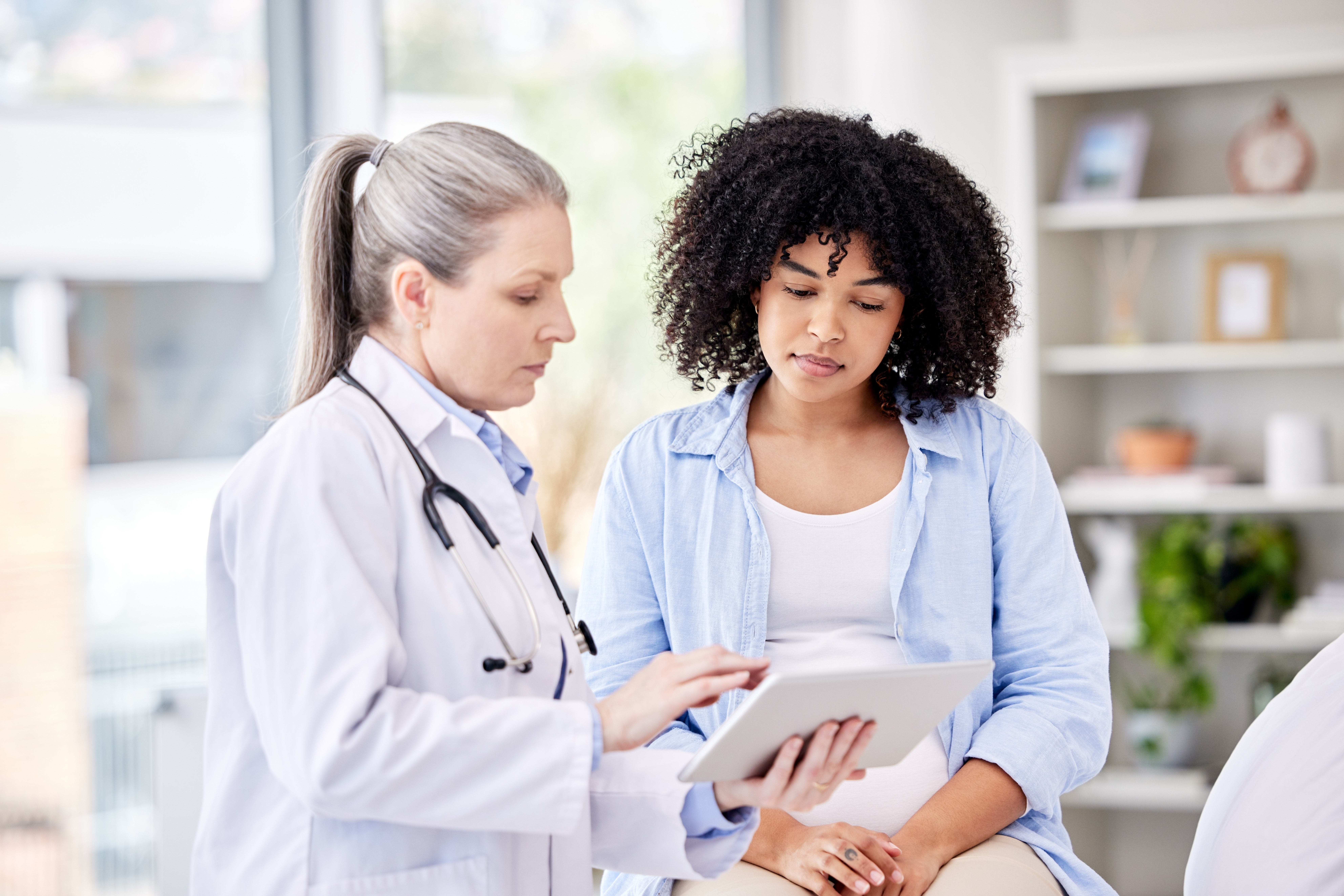 A doctor shows a tablet to a young woman patient sitting beside her in a medical office; the two appear to be discussing health information together.