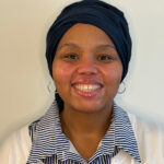Karyn Stewart smiling, wearing a dark headwrap, hoop earrings, and a white cardigan over a blue and white striped button-down shirt.