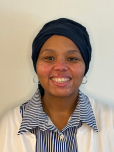 Karyn Stewart smiling, wearing a dark headwrap, hoop earrings, and a white cardigan over a blue and white striped button-down shirt.